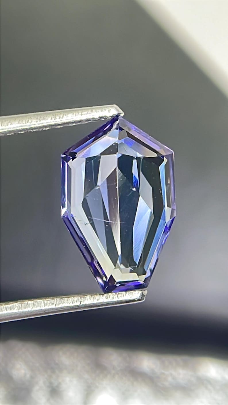 May include: A faceted, purple gemstone with a hexagonal shape, held by tweezers against a grey background.