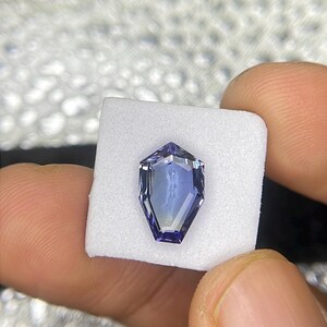 May include: A purple gemstone with a faceted, octagonal shape. The gemstone is set in a white, square-shaped display box.