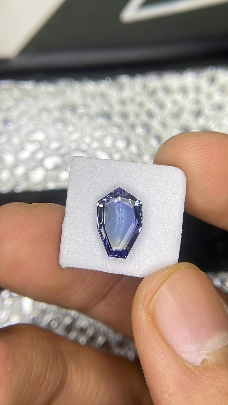 May include: A purple gemstone with a faceted, octagonal shape. The gemstone is set in a white, square-shaped display box.