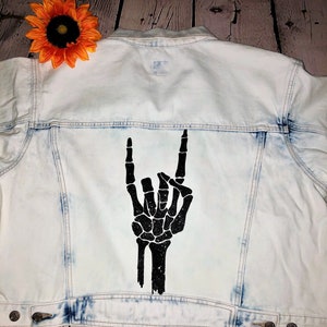 May include: Light blue denim jacket with a black skeleton hand making the rock and roll hand gesture on the back.
