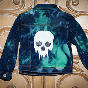 May include: A blue denim jacket with a white skull design on the back. The skull is dripping with white paint and has black eyes. The jacket has a tie-dye pattern in shades of blue and green.