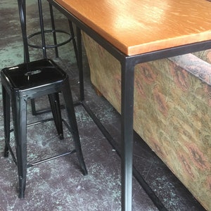 May include: A long, rectangular wooden table with a black metal frame. The table is sitting on a concrete floor and has two black metal stools next to it.