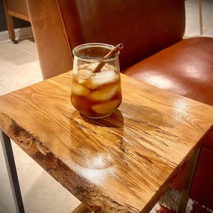 May include: A glass of iced tea with a cinnamon stick, sitting on a wooden end table with a live edge top. The table has a natural wood finish and is supported by a metal frame.