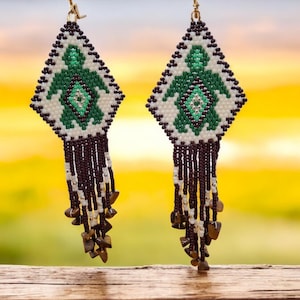 Peut inclure: Une paire de longues boucles d'oreilles en perles avec un motif géométrique représentant une tortue verte. Les boucles d'oreilles sont composées de perles brunes, blanches et argentées, et d'accents de pierres précieuses brunes.