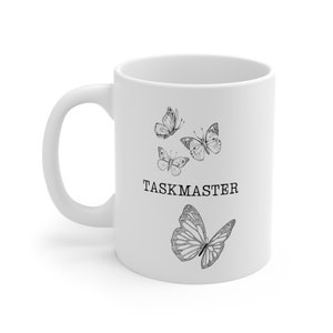 Könnte beinhalten: Weiße Keramiktasse mit einer schwarzen Schmetterlingsillustration und dem Wort "TASKMASTER". Die Tasse hat einen gebogenen Henkel und ist für heiße oder kalte Getränke geeignet. Das Design umfasst drei kleinere Schmetterlinge über dem Text und einen großen Schmetterling darunter.