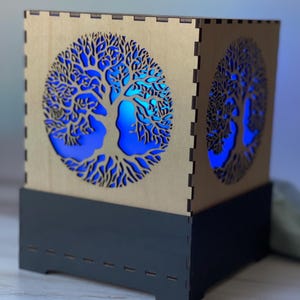 Könnte beinhalten: Eine Holzlampe mit einem Lebensbaum-Motiv. Die Lampe ist mit blauem Licht beleuchtet, was eine warme und einladende Atmosphäre schafft.