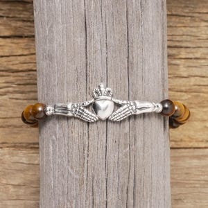 May include: A brown tiger's eye beaded bracelet with a silver Celtic knot charm featuring two clasped hands holding a heart with a crown on top.