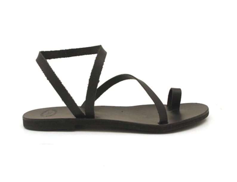 May include: Black leather sandal with a toe loop and ankle strap. The sandal features a flat sole and a minimalist design, perfect for warm weather. The sandal is shown on a white background.