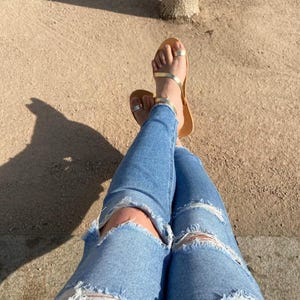 May include: Light blue ripped jeans and gold sandals with a toe ring. The sandals have a thin, gold strap across the foot and a brown sole. The jeans have distressed details at the knees.