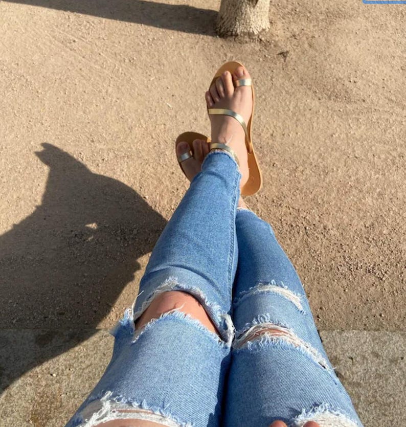 May include: Light blue ripped jeans and gold sandals with a toe ring. The sandals have a thin, gold strap across the foot and a brown sole. The jeans have distressed details at the knees.