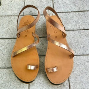 May include: A pair of tan leather sandals with metallic rose gold straps. The sandals have a toe loop and ankle strap design. The soles are a light brown color, and the size 37 is stamped on the footbed. The sandals are displayed on a gray tiled surface.