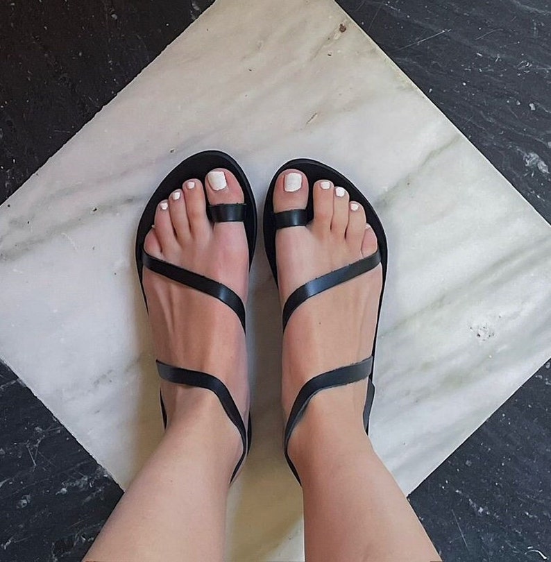 May include: Black leather sandals with a toe loop and multiple straps. The sandals are worn on a marble floor with a black and white geometric pattern. The person's toes are painted white.