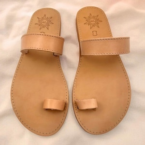 Könnte beinhalten: Ein Paar hellbraune Ledersandalen mit einem breiten Riemen über dem Mittelfuß und einer separaten Schlaufe für den großen Zeh. Das Fußbett ist mit einem Sonnensymbol und dem Schriftzug "Hellas" in Fersennähe sowie sichtbaren Nähten versehen.
