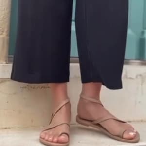 May include: Tan-colored sandals with a toe loop and ankle strap. The sandals have a flat sole and are worn with black cropped pants. The sandals are made of a soft material and are suitable for warm weather.