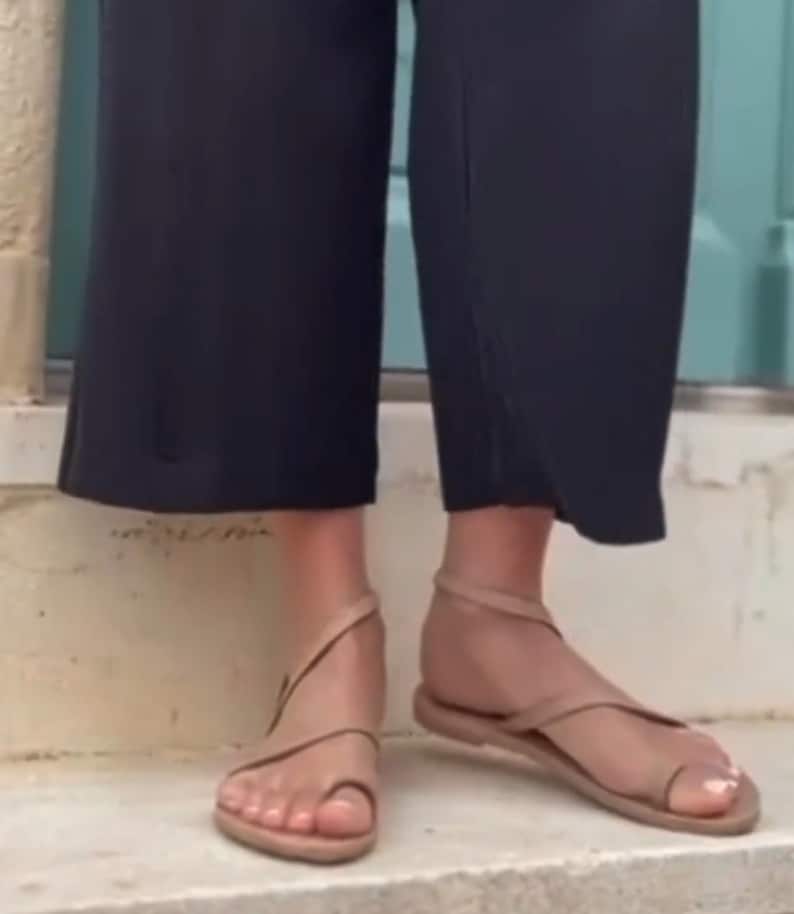 May include: Tan-colored sandals with a toe loop and ankle strap. The sandals have a flat sole and are worn with black cropped pants. The sandals are made of a soft material and are suitable for warm weather.