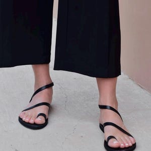 May include: Black leather sandals with a toe loop and ankle strap. The minimalist design features thin straps that wrap around the foot. The sandals are worn with black wide-leg pants, creating a sleek, monochromatic look.