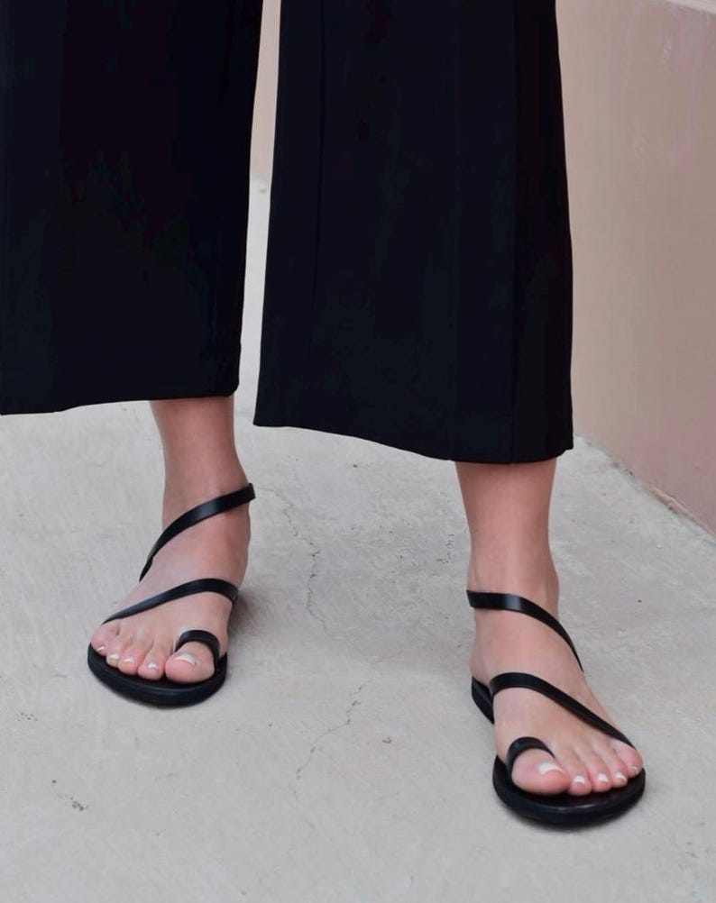 May include: Black leather sandals with a toe loop and ankle strap. The minimalist design features thin straps that wrap around the foot. The sandals are worn with black wide-leg pants, creating a sleek, monochromatic look.
