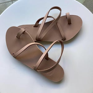 May include: A pair of light brown leather sandals with a toe loop and multiple straps. The sandals are flat and appear to be handmade. The sandals are displayed on a white surface.
