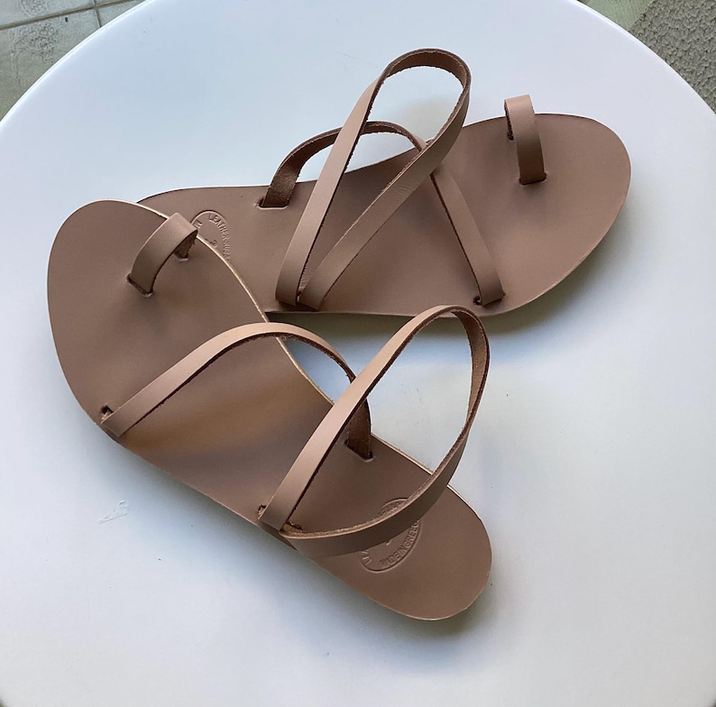 May include: A pair of light brown leather sandals with a toe loop and multiple straps. The sandals are flat and appear to be handmade. The sandals are displayed on a white surface.