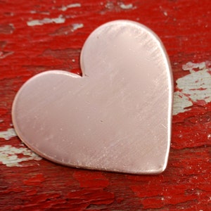 May include: A heart-shaped piece of copper metal, with a smooth, polished surface, is sitting on a red, weathered wooden surface.