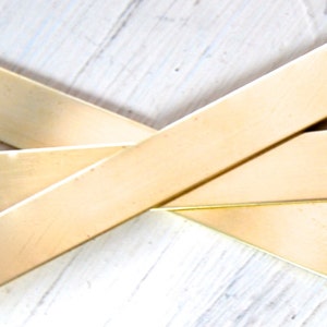 May include: Four rectangular, gold-colored metal bars are arranged diagonally across a light-colored wooden surface. The bars have a slightly brushed texture and a warm, metallic sheen. The composition suggests a focus on geometric shapes and materials.