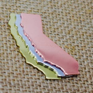 May include: Three layered metal cutouts of the state of California. The top layer is copper, the middle layer is silver, and the bottom layer is gold.