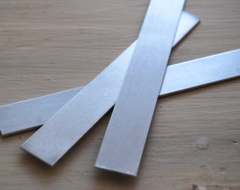 May include: Close-up of three brushed metal strips, each rectangular and silver in color. The strips are arranged diagonally across a light brown wooden surface, with their edges slightly overlapping. The metal has a matte finish.
