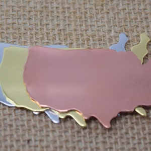 May include: Three metal outlines of the United States of America, stacked on top of each other. The top outline is silver, the middle outline is gold, and the bottom outline is copper.