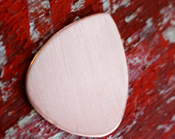 May include: A blank, copper guitar pick with a rounded, triangular shape. The pick is a light pink color and has a smooth, polished surface.