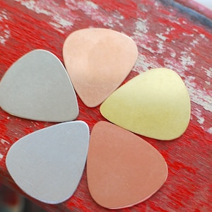 May include: Five guitar picks made of different metals. The picks are arranged in a flower shape. The picks are copper, brass, silver, and two shades of bronze.