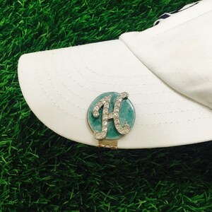 May include: A white baseball cap featuring a circular golf ball marker attached to the brim. The marker has a teal marbled background with a silver-toned letter H encrusted with clear rhinestones centered on the surface of the circular disc.