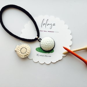 May include: A golf ball marker necklace with a black cord, a gold magnetic ball marker, and two golf tees. The golf ball marker is white with a black border. The text "fuluya" and "GOLF BALL MARKER" are on a white scalloped card.