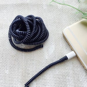 May include: A navy blue and white striped phone charging cable is connected to a gold smartphone. The cable is coiled and has a protective, spring-like design. The background is a textured, off-white surface.