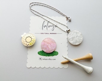 May include: A set of golf ball markers with a white, pink, and gold design. The markers are attached to a silver chain necklace. The image also includes two golf tees, one white and one brown.