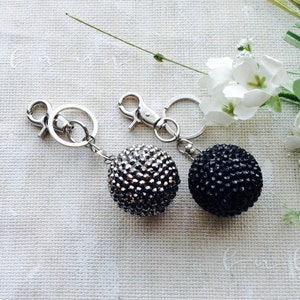May include: Two spherical keychains featuring a textured surface covered in small, reflective beads. One sphere is silver and the other is black. Each keychain includes a silver-toned metal lobster claw clasp and a circular split ring for attachment.