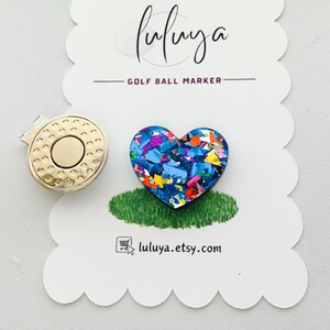 May include: A heart-shaped golf ball marker with a blue base and colorful confetti-like glitter. The marker is attached to a gold-colored magnetic clip.