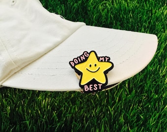May include: A yellow star-shaped pin with a smiling face and the words "DOING MY BEST" in pink lettering, attached to a white hat. The pin is set against a backdrop of green grass.