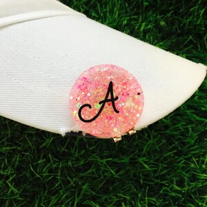May include: A pink and white glitter golf ball marker with the letter "A" in black. The marker is attached to a white golf hat.