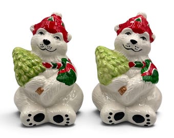 May include: A pair of ceramic polar bear salt and pepper shakers. Each white bear wears a red hat with white trim and a red and green scarf, while holding a light green textured Christmas tree. The bears have black painted noses, mouths, and paw pads.