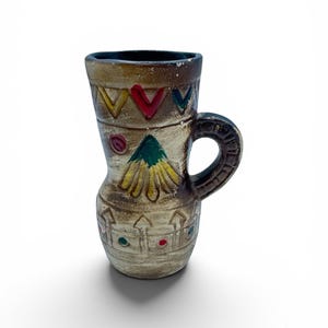 Könnte beinhalten: Ein brauner Keramikbecher mit ausgestelltem Rand und einem einzelnen Henkel. Die Außenseite zeigt handgemalte Designs, darunter gelbe, rote und grüne Zickzackmuster, ein zentrales grün-gelbes Dreiecksmotiv und ein unteres Band mit zaunartigen Formen.