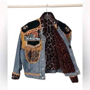 May include: A denim jacket with elaborate embellishments. The jacket features patches of dark green velvet, gold fringe, and intricate embroidery. The interior lining has a leopard print design. The jacket is hanging on a metal hanger.