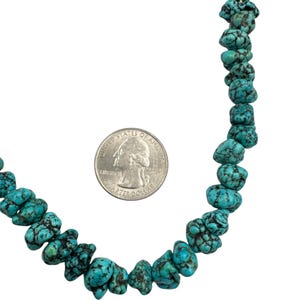 Puede incluir: Un collar hecho de cuentas de turquesa irregulares y pulidas con vetas de matriz oscura. Una moneda de veinticinco centavos de los Estados Unidos se coloca junto a las cuentas para proporcionar una referencia visual del tama&ntilde;o de las piedras.
