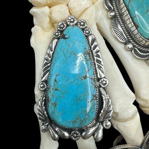 Puede incluir: Primer plano de joyas de turquesa con engastes de plata. Las piedras de turquesa son de un azul vibrante con vetas marrones. Los engastes de plata tienen intrincados diseños florales y de hojas. Las joyas se exhiben sobre un fondo blanco.