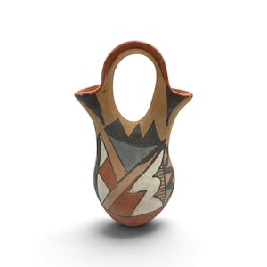 Pode incluir: Um vaso de cerâmica com uma alça central arqueada e duas aberturas laterais. A superfície externa é decorada com padrões geométricos em tons de terracota, preto, branco e cinza, aplicados sobre uma base de argila clara.