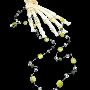 Puede incluir: Un collar con un colgante de mano de hueso. El collar presenta una cadena plateada con cuentas de piedras preciosas amarillas, verdes y transparentes. La mano de hueso es blanquecina y detallada, con huesos de los dedos visibles. El fondo es negro.