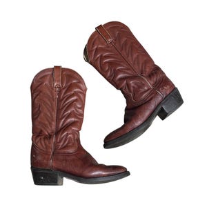 Könnte beinhalten: Ein Paar braune Leder-Cowboystiefel mit aufwendigen Steppmustern auf den hohen Schäften, Zuglaschen am oberen Rand, abgerundeten Zehenpartien und Blockabsätzen. Die Stiefel haben eine abgenutzte Textur und eine dunkelbraune Sohle mit einem kleinen Stern.