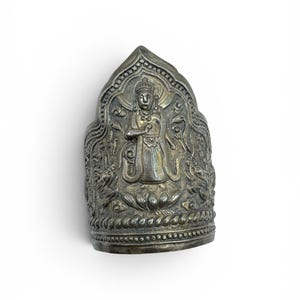 Puede incluir: Un artefacto religioso plateado y ornamentado con un relieve detallado de una figura coronada, rodeada de intrincados patrones. El objeto tiene una base curva y una parte superior puntiaguda, probablemente una pieza decorativa o ceremonial.