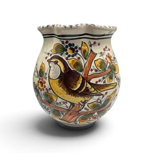 May include: A ceramic vase with a cream-colored base featuring a hand-painted design of a brown and yellow bird perched on a branch. The artwork includes green leaves, small red berries, and orange flowers. The vase has a scalloped, wavy rim.