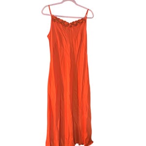 Op de afbeelding: Een oranje slipdress met spaghettibandjes en een kanten halslijn. De jurk hangt aan een roze hanger tegen een witte achtergrond. De jurk is effen van kleur en lijkt van een zijdeachtig materiaal te zijn.