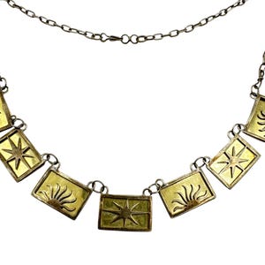 Puede incluir: Un collar plateado con colgantes rectangulares con diseños de sol y estrella en oro y amarillo. El collar tiene una cadena delicada y un cierre sencillo, adecuado para el uso diario.